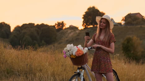 Beautiful-blonde-in-a-hat-with-a-bike-looking-at-the-mobile-phone-screen-and-a-basket-on-the-handlebar-with-flowers